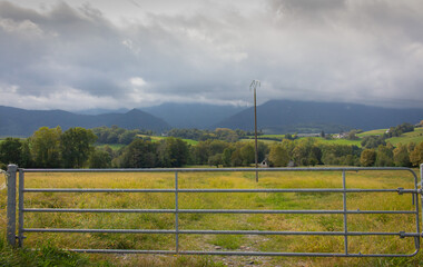 Fototapeta premium Closed iron fence in the field. Meadow and mountauns on background. Countryside concept. Cloudy day in rural pasture. Electric pole in field behind fence. Summer nature concept. 