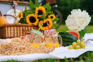 Summer picnic concept on sunny day with watermelon, fruit, bouquet hydrangea and sunflowers flowers. Picnic basket on grass with food and refreshing summer drink on white knit blanket. Selective focus