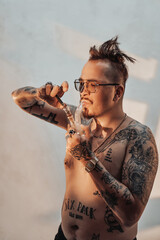 Shirtless man with tattooed body and freaky haircut lighting cannabis in the bong. Cannabis legalization concept