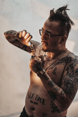 Shirtless man with tattooed body and freaky haircut lighting cannabis in the bong. Cannabis legalization concept
