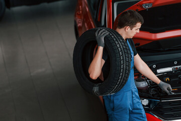 Worker in black and blue uniform holds car wheel and walks with in near red automobile