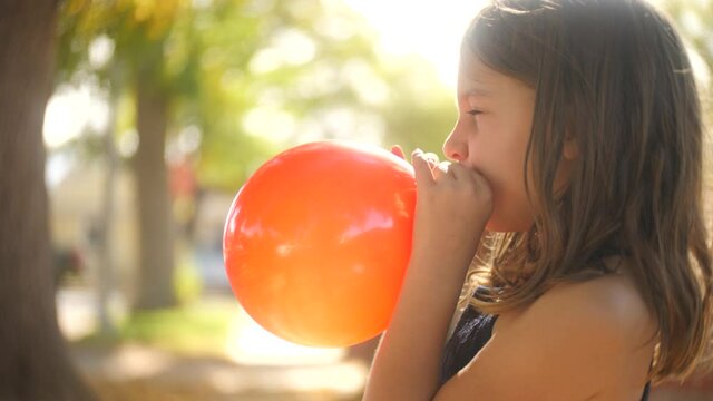 Cute 7 Year Old Girl Child Blowing Up Inflatable Orange Balloon In Summer Street. Shallow DOF, Slow Motion.