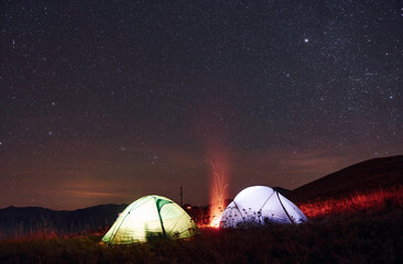 Two iluminated tents with campfire under stars at mountains at night