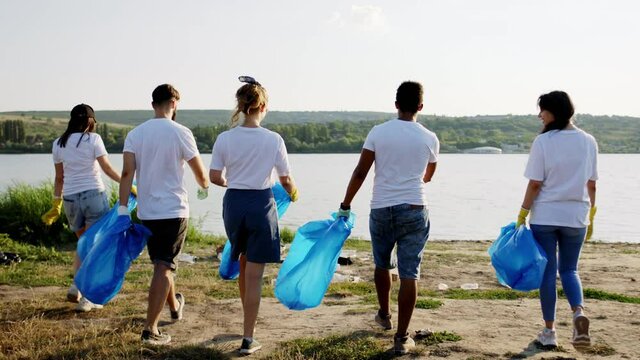On the beach side beside the lake group of multiracial volunteers walking down the lake holding big plastic bags to collecting the rubbish they picking up the rubbish and collecting on the bags. 4k