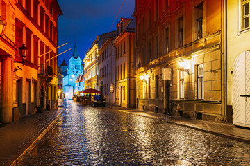 Fototapeta na wymiar Riga, Latvia. Evening View Of Pils Street With Ancient Architecture In Bright Yellow Illumination Under Summer Blue Sky. Our Lady Of Sorrows Or Virgin Of Anguish Church In Distance