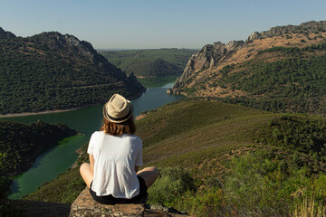 Fototapeta na wymiar Attractive young female with a hat sitting on top of a rock in a viewpoint witnessing a river flowing between the mountains in Monfrague, Spain.