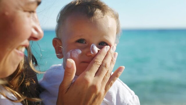 Close up of young mother is applying a protective sunscreen or sunblock lotion on her little toddler son face to take care of his delicate skin on a seaside beach during family holidays vacation.