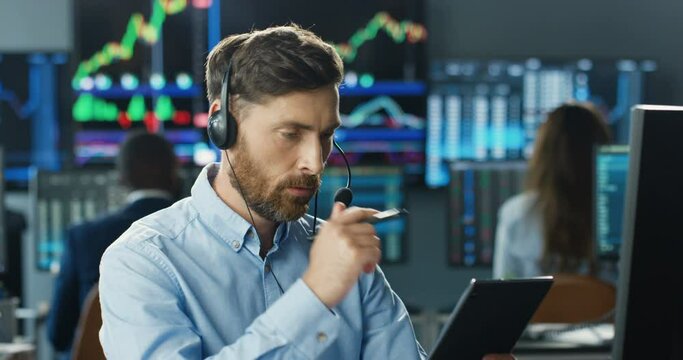 Handsome young Caucasian man trader in headset using tablet device while thinking and working in trading office. male broker putting bets, selling and buying online.