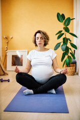 Middle aged fit focused pregnant yogi woman sitting in lotus position at home and meditating.