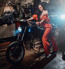 Attractive sexual young brunette in orange jumpsuit staying with a modern motorcycle in the car service