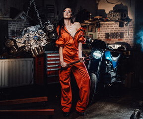 Fototapeta na wymiar Brave and very attractive looking young woman posing with a big wrench, surrounded by motorcycle details and with a black motorbike on the background