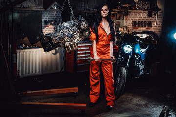 Brave and very sexy looking young woman in safety googles, posing with a big wrench, surrounded by motorcycle details and with a motorbike on the background