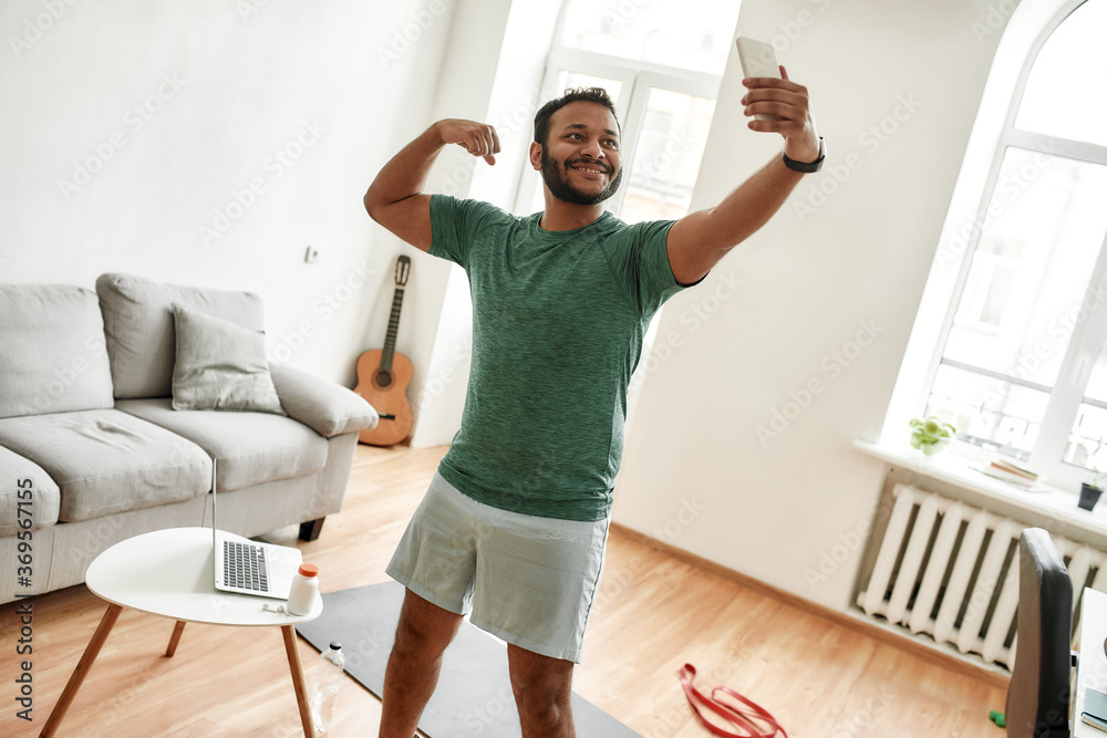 Wall mural Be physically fit. Excited bearded active man holding smartphone, showing biceps while taking a selfie during morning workout at home. Fitness, motivation concept