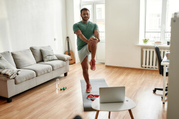 Full length shot of young active man watching online video training on laptop, exercising during morning workout at home. Sport, healthy lifestyle