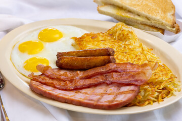 A view of a breakfast plate, featuring sunny side up eggs, ham, bacon, sausage, hash browns and toast, in a restaurant or kitchen setting.