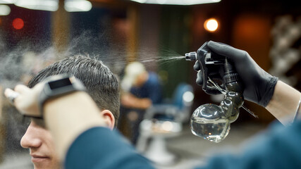 Hairstyling. Close up detail shot of a professional barber spraying hair of young guy visiting modern barbershop