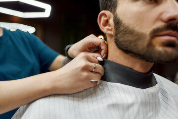 Ready to work. Close up shot of a handsome bearded man sitting in barbershop chair. Barber girl or female hairdresser preparing client for a haircut