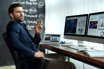 Fototapeta na wymiar Young confident businessman in wireless earphones talking with client or partner, discussing sales report while working with statistical data in the modern office