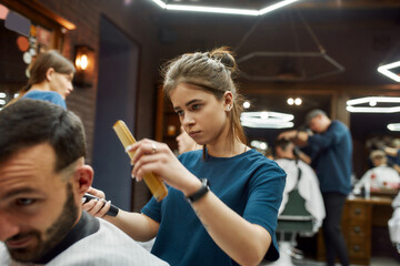 Close up shot of a professional barber girl or female hairdresser working with hair clipper, making trendy haircut for a handsome bearded man