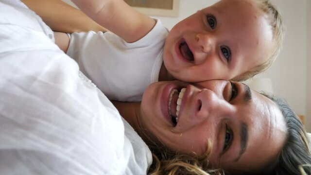 Authentic close up shot of an young happy smiling mother is enjoying to have fun with her newborn baby in a bedroom just woke up in morning.