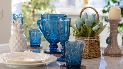 Light dining room interior with a large window. Marble table with oak base. Serving with white dishes and a blue accent. Blue glasses, flowers.