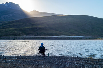 Camping area on a lake in mountainous terrain. A woman with a camp chair and a tea thermos