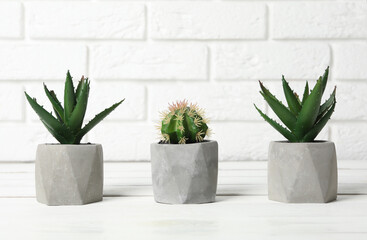 Beautiful artificial plants in flower pots on white wooden table near brick wall