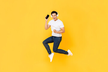 Fototapeta na wymiar Happy young Asian man wearing headphone listening to music from mobile phone and jumping isolated on yellow background