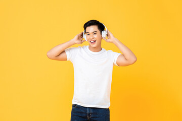 Happy young handsome Asian man listening to streaming music on wireless headphones isolated on yellow studio background