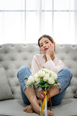 Beautiful charming Asian woman laying on white sofa and holding surprise white rose bouquet from her boyfriend at living room in the morning. Happiness relaxation mood concept.