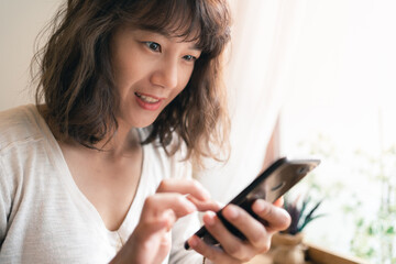 Close up of young Asian woman using smartphone chatting and searching information with blurry smiley face background. Technology business and social distancing concept