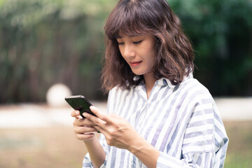 Young Asian woman using smartphone chatting and searching information. A happy female with smiley face in outdoor park. technology business and social distancing concept
