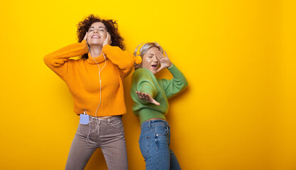 Obraz premium Charming two women dancing on a yellow studio wall with free space listening to music through earphones
