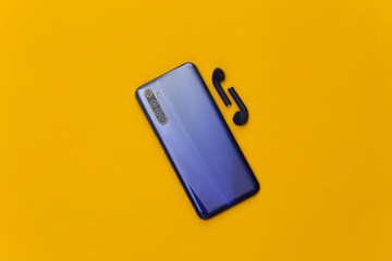 Modern smartphone with wireless earphones on yellow background. Top view. Flat lay