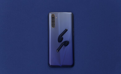 Modern smartphone with wireless earphones on classic blue background. Color 2020. Top view. Flat lay