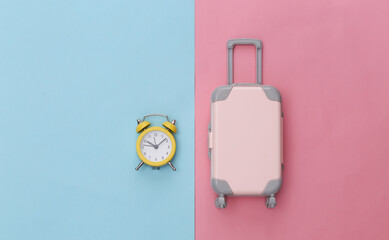 Mini luggage and alarm clock on pink blue pastel background. Time to travel. Top view