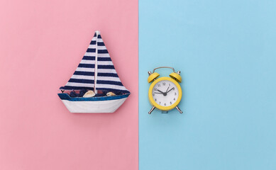 Mini alarm clock and mini sailboat on pink blue pastel background. Time to travel. Top view