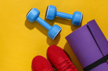 Red sports shoes, dumbbells and yoga mat on a yellow bright background with deep shadow. Fitness composition. Flat lay. Top view