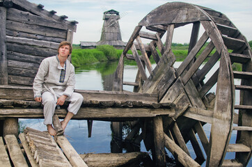 young man in old linen clothes sits on the pier of an old water mill