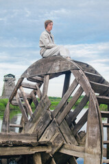 young man in old linen clothes sits on the wheel of an old water mill