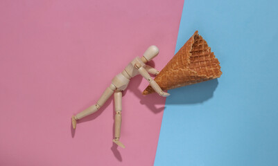 Wooden puppet holding Ice cream waffle cone on pink blue background. Top view
