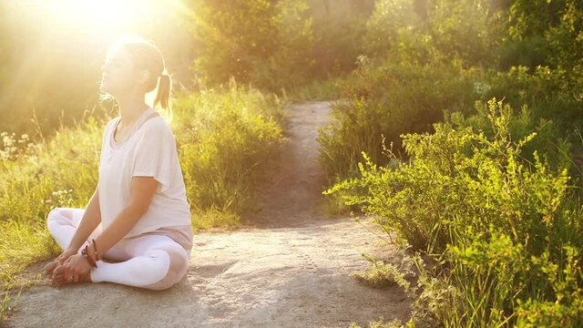 Calmly young yogini woman sitting Bound Angle Pose and raising head to sun outside in nature evening on background of bright sunray. Meditative girl practicing yoga exercises in alone outdoors sunset.