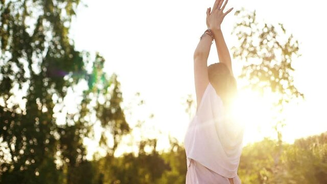 Rear view of unrecognizable young woman meditating holding hands together in Namaste mudra posture. Relaxed female meditating raising hands outside in park evening on background of soft sunbeams.