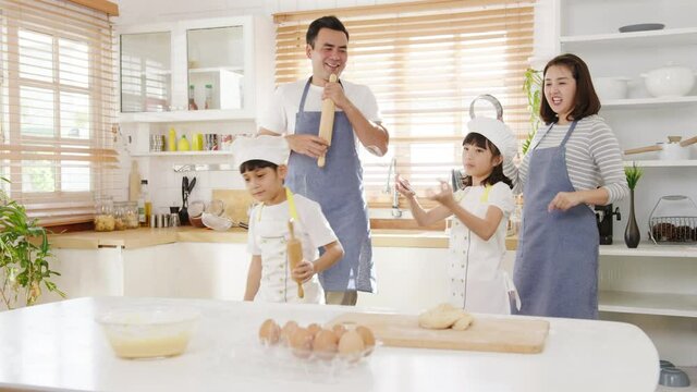 Happy young Asian family with preschool kids have fun listen to music and dancing while cooking baking pastry for breakfast meal in kitchen home in morning. Doing bakery knead dough and bake cookies.