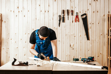 A young brunette man builder handles a wooden bar with a black jack plane in the workshop, in the...