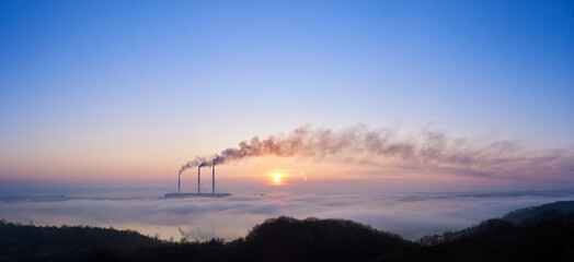 Fototapeta na wymiar Panoramic view of three smoking stacks of thermal power station on the horizon taken from the hill, pipes are in evening fog on blue sky, copy space. Concept of ecology and environmental pollution.