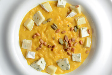 Creamy pumpkin soup topped with pumpkin seeds and cheese in a white bowl on a wooden table with plaid tablecloth.