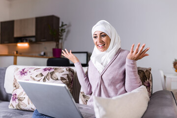 Young Muslim woman having video call via laptop at home. Happy smiling Muslim woman sitting on sofa, couch and using laptop at living room at home, learning language, video calling.