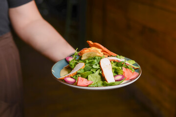 Fresh vegetable salad with chicken meat and tomatoes. Caesar salad. Served in a blue plate. In hand of a waiter.