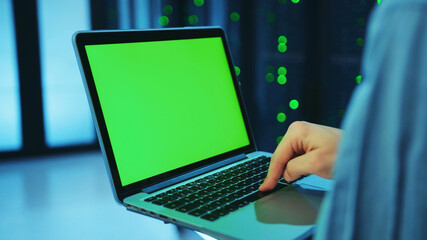 Over the shoulder IT male administrator keyboarding on laptop chrome key green screen, running server diagnostics in database cabinet. Close-up worker with mock-up notebook at data center.